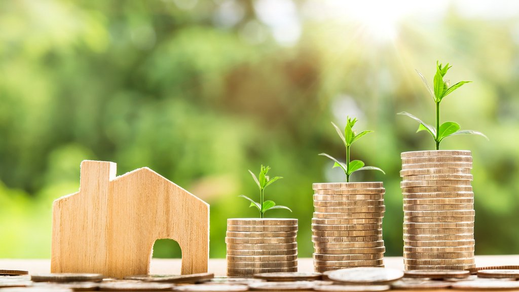 Symbolisches Holzhaus neben gestapelten Münzen mit wachsenden Pflänzchen als Zeichen für Immobilieninvestitionen und finanzielles Wachstum