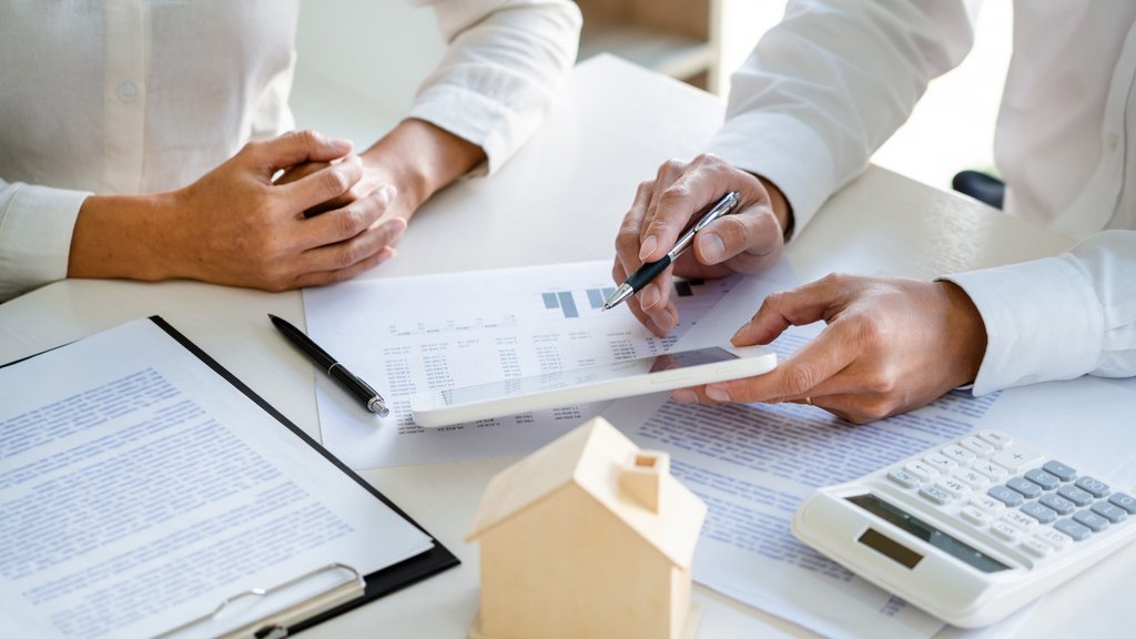 Zwei Personen besprechen Finanzdokumente mit Taschenrechner, Tablet und Miniaturhausmodell auf dem Tisch